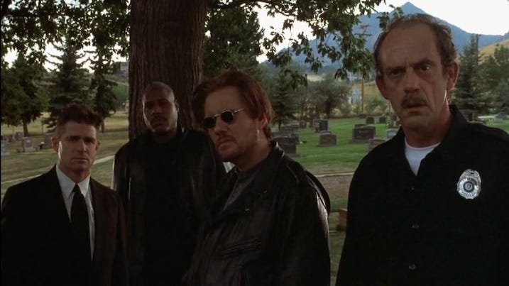 Funeral Crew: Four men, including Christopher Lloyd, stand in a cemetery dressed in black, eyeing someone offscreen with a mix of suspicion and unease.