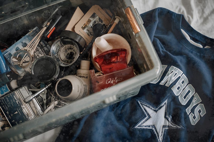 A plastic bin filled with small keepsakes — toiletries, tools, and mementos — sits beside a folded Cowboys sweatshirt on a bed A plastic bin filled with small keepsakes — toiletries, tools, and mementos — sits beside a folded Cowboys sweatshirt on a bed