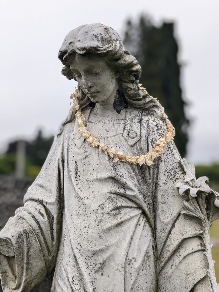 Bess provided several images of her time in cemeteries. The first image shows a bramble of ripening blackberries. The second image shows Bess Lovejoy, with dark hair and wearing a black top and green skirt, gazing upon an aging headstone. The third image features a statue of a woman, face looking downward, and is adorned with a flower necklace. The final photo is a cluster of headstones, one spiking higher than the others, but all showing decades of age.