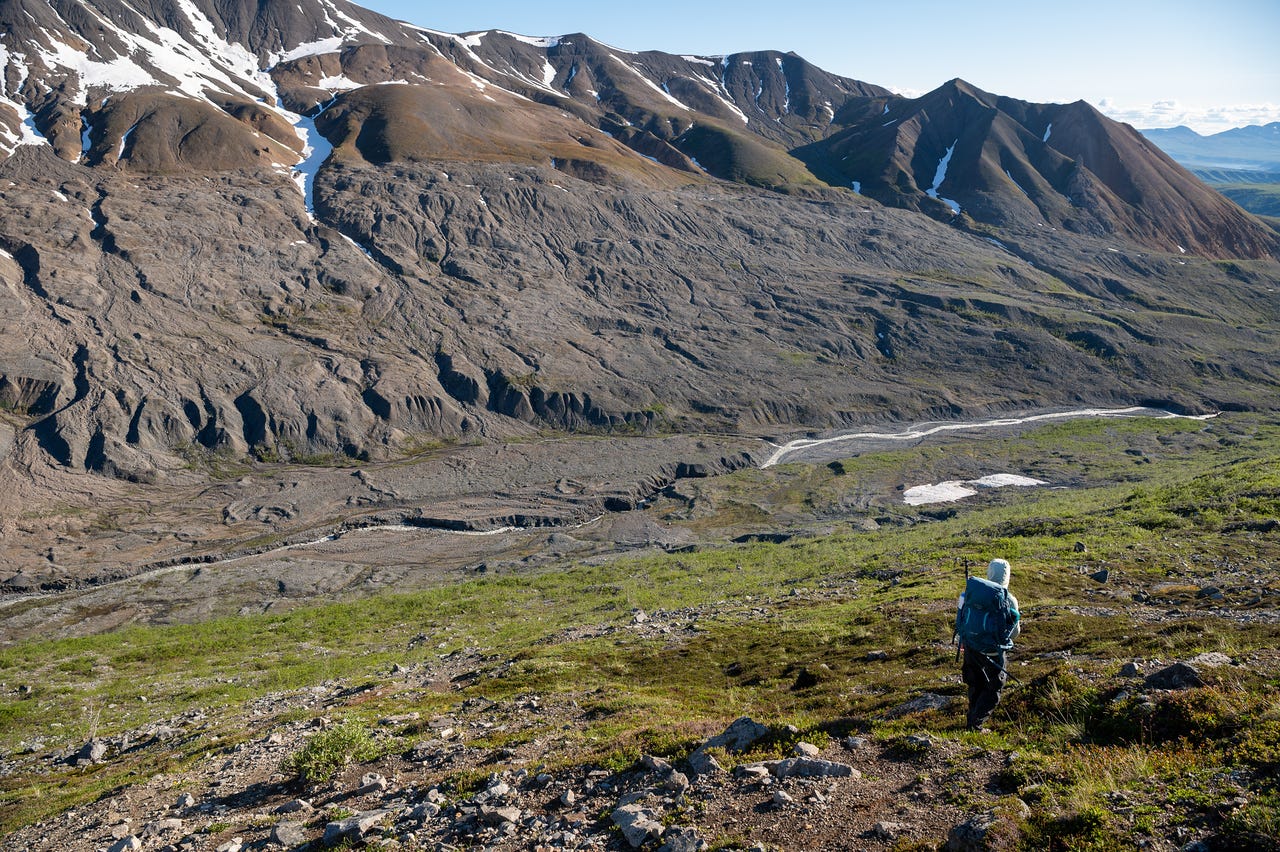 A small hooded figure with a pack walks down a green tundra slope toward a wide valley of grey rubble and braided meltwater channels. Across the valley, a long mountainside is marked by a clear horizontal line where scoured grey rock below meets vegetated slopes above, snow patches lingering along the crest under a bright sky.