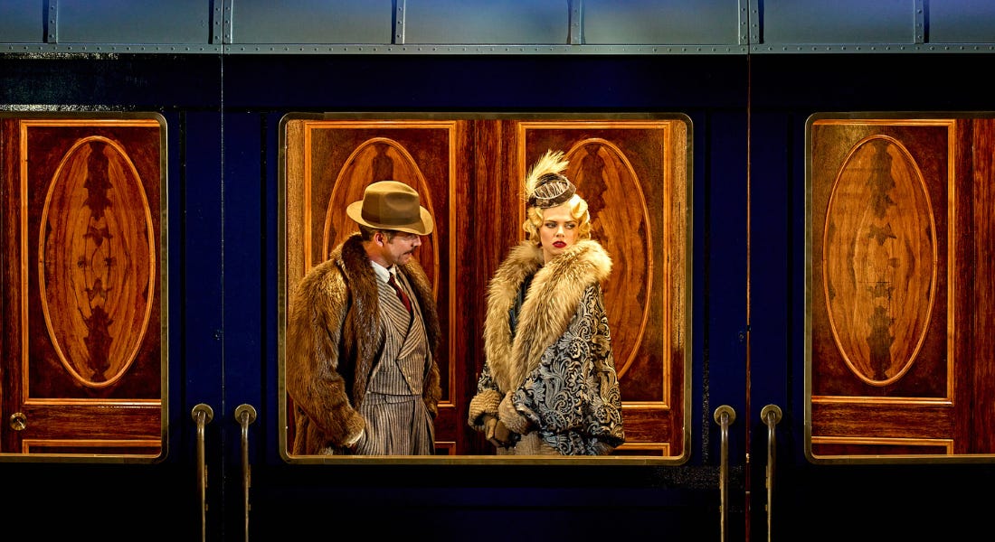 A blue train carriage with a man in a fur coat and suit and a woman wearing a grey dress stood inside of it