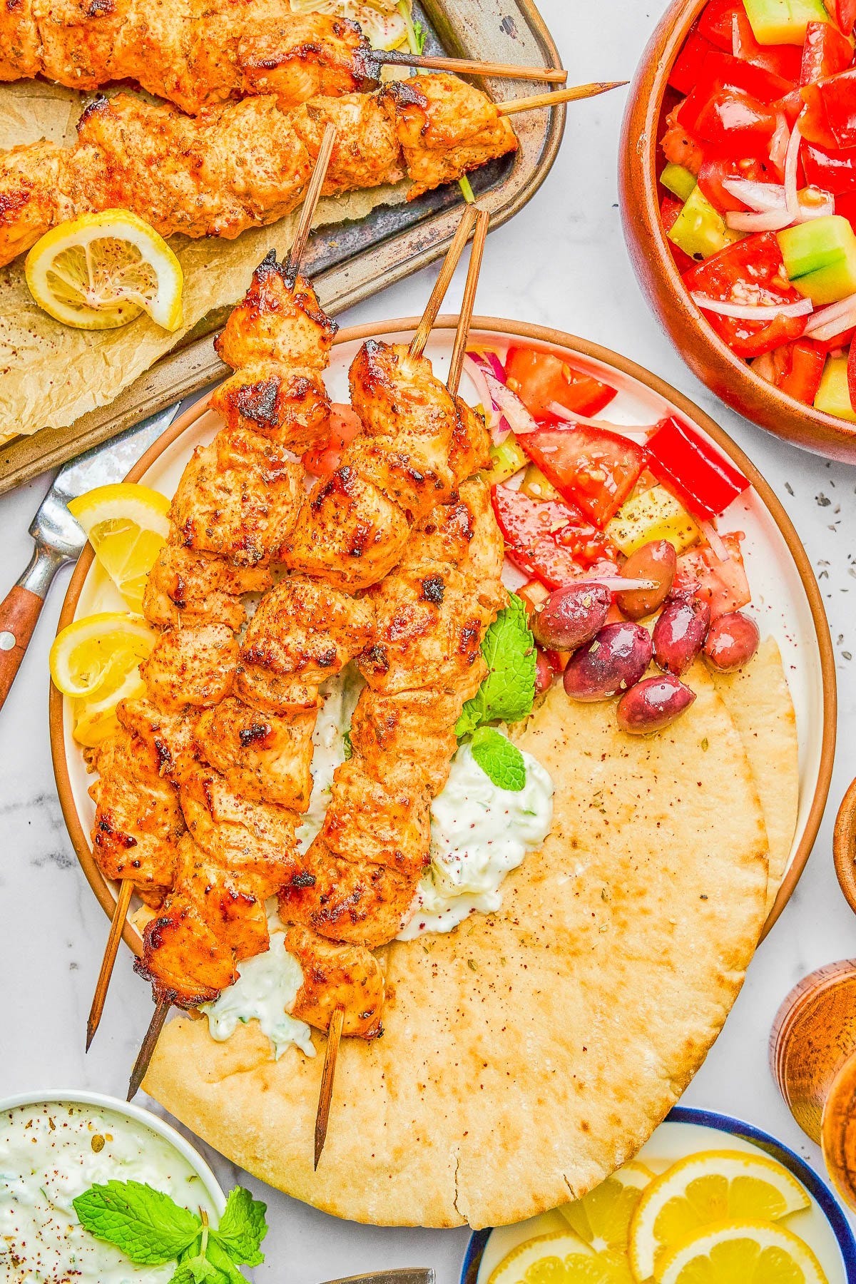 Grilled chicken skewers on pita bread with tzatziki, lemon wedges, olives, and a tomato-cucumber salad, served on a white plate.