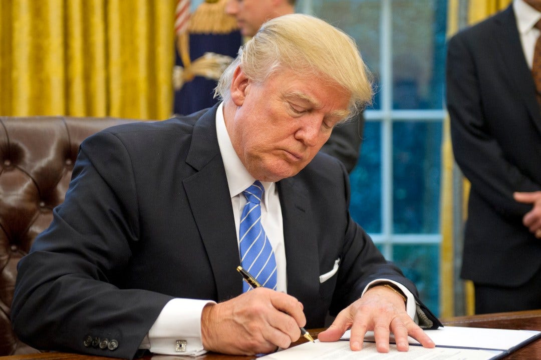 Donald Trump seated at wooden desk signing document with pen, wearing dark suit white shirt blue tie, focused expression, gold curtains behind, another suited man standing nearby with hands clasped, leather chairs visible.