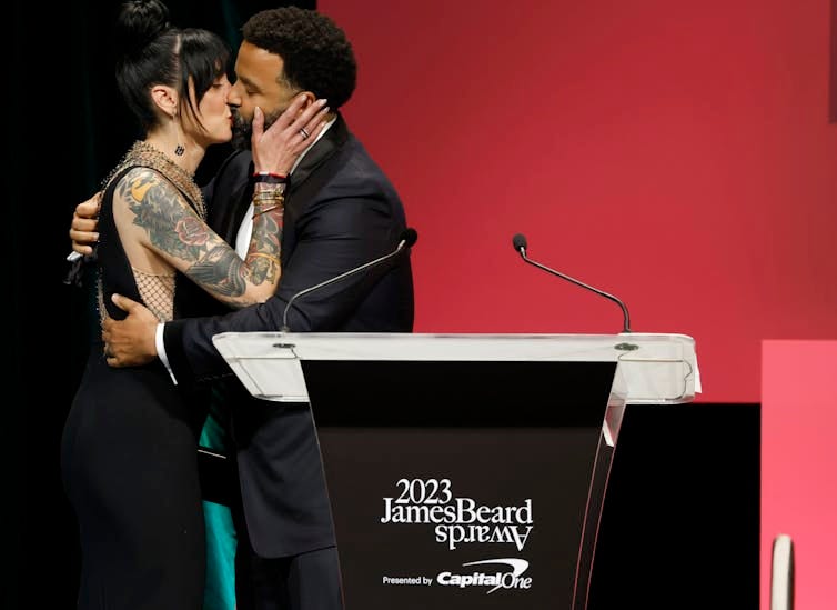 Man and women in formalwear kiss on a stage behind a podium that says '2023 James Beard Awards'