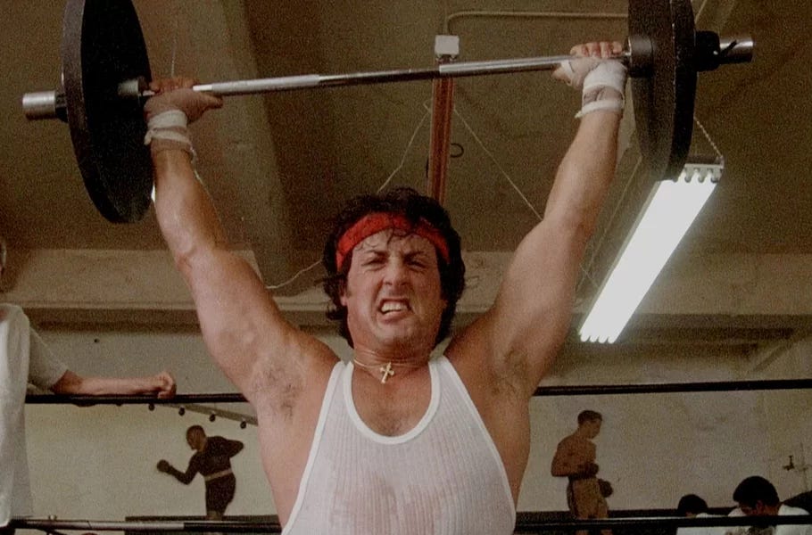 Photo of Sylvester Stallone as Rocky Balboa lifting weights in the movie Rocky (1976)