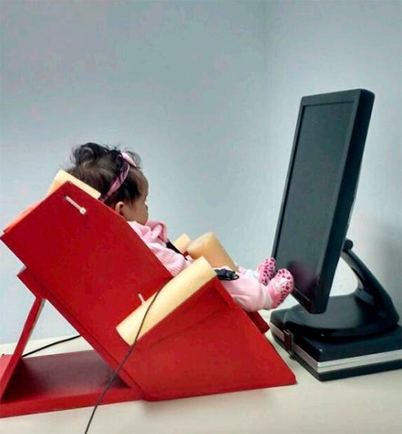 Bebê usando laço e roupa rosa sentado em uma cadeira vermelha com braços amarelos, tendo sensores conectados nas mãos, posicionada em frente ao monitor de vídeo Bebê usando laço e roupa rosa sentado em uma cadeira vermelha com braços amarelos, tendo sensores conectados nas mãos, posicionada em frente ao monitor de vídeo