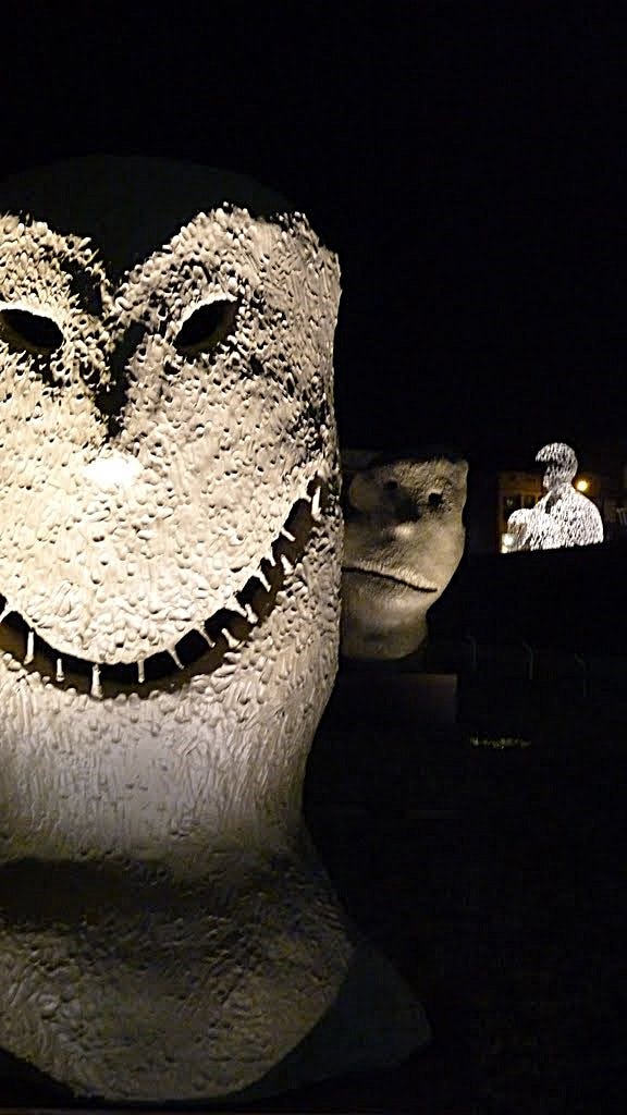 Moonrise sculptures at night in the Pappajohn Sculpture Garden in Des Moines. They're big, textured faces. The one in front is spooky with big teeth. The one behind it looks like it's peeking around the spooky face and looks concerned. Nomad, the man made of letters, is lit up in the background.