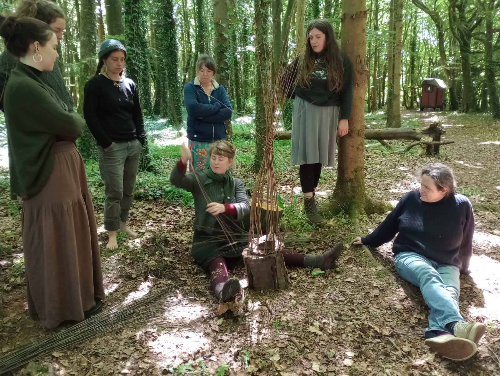 Rosemary sits on the ground, the beginnings of a basket in her hands. She is speaking and we are gathered around her, listening, watching. Five of us stand, one leans against a tree, one is seated on the ground. Behind us, a tall fence of ivy and moss wrapped tree trunks.