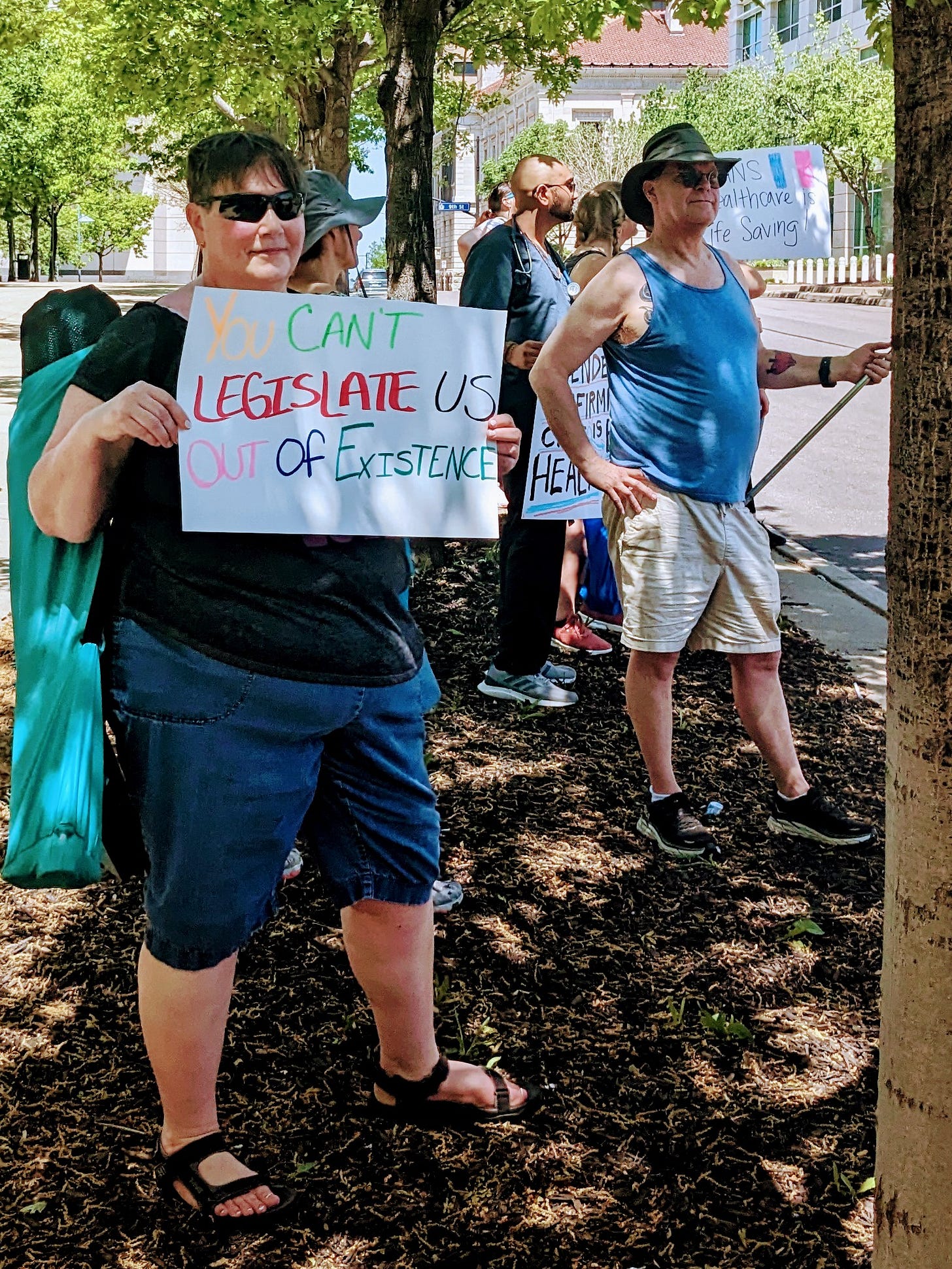 A group of protestors holding signs that read "You Can't Legislate Us Out of Existance," "Gender-Affirming Care is HEALTHCARE," and "Trans Healthcare is LIFE-SAVING."