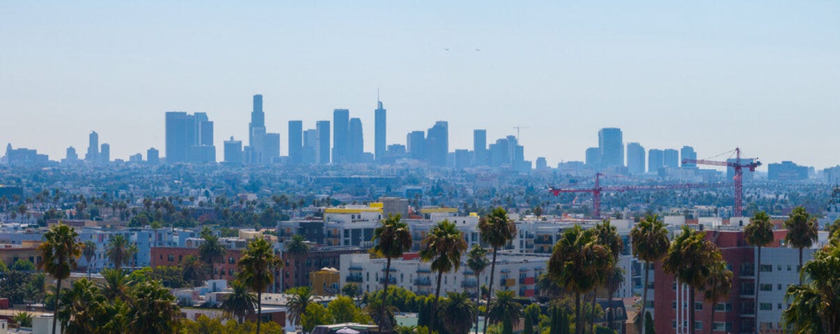 The image features Los Angeles skyline with downtown skyscrapers, palm trees, and mid rise buildings under a clear blue sky with visible construction cranes.