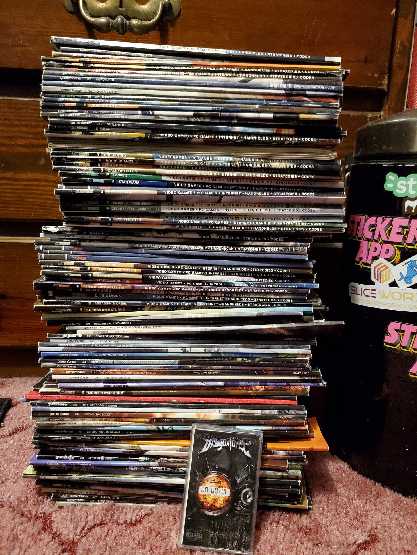 A tall stack of Game Informer magazines. They are unorganized and unevenly lined up. Next to them is a Dragonforce cassette (Inhuman Rampage) and a trashcan to give a sense of scale. The cassette is dwarfed by the height of the stack.