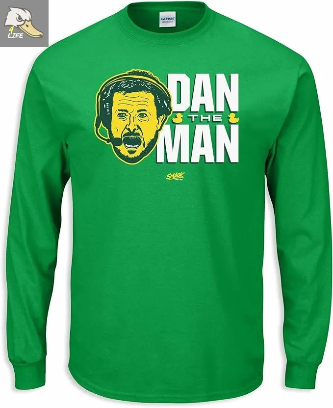 Green long-sleeve Oregon football shirt featuring a bold “Dan The Man” design with a stylized illustration of the Ducks’ head coach wearing a headset, white and yellow text, and a small Ducks4.Life logo in the corner. Deals on seats, fan gear, and premium Oregon Football coverage at www.Ducks4.Life 