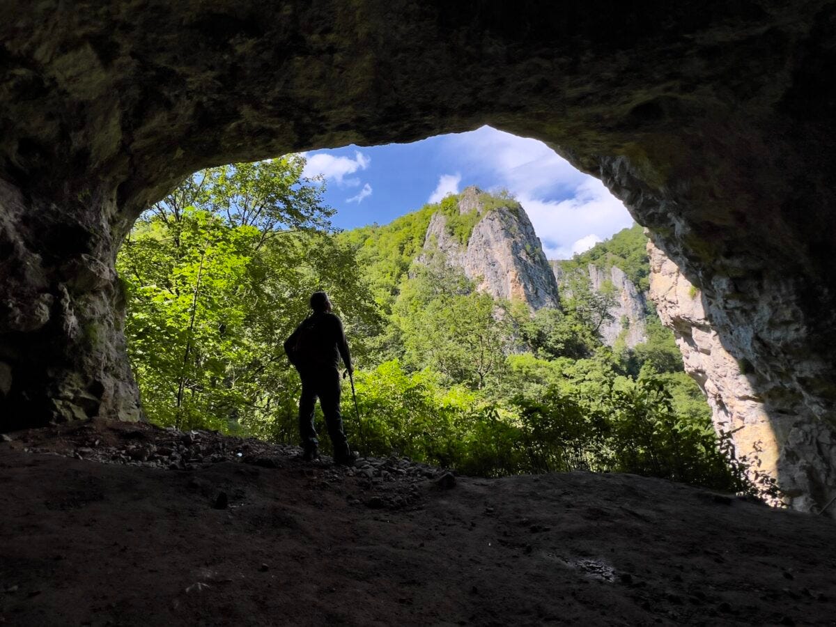 Imagem mostra a silhueta de um pesquisador parado em frente à entrada da caverna Pestera Mare. Ao fundo é possível ver a paisagem verdejante do cânion Garganta do Varghis - Foto: Herton Escobar / USP Imagens