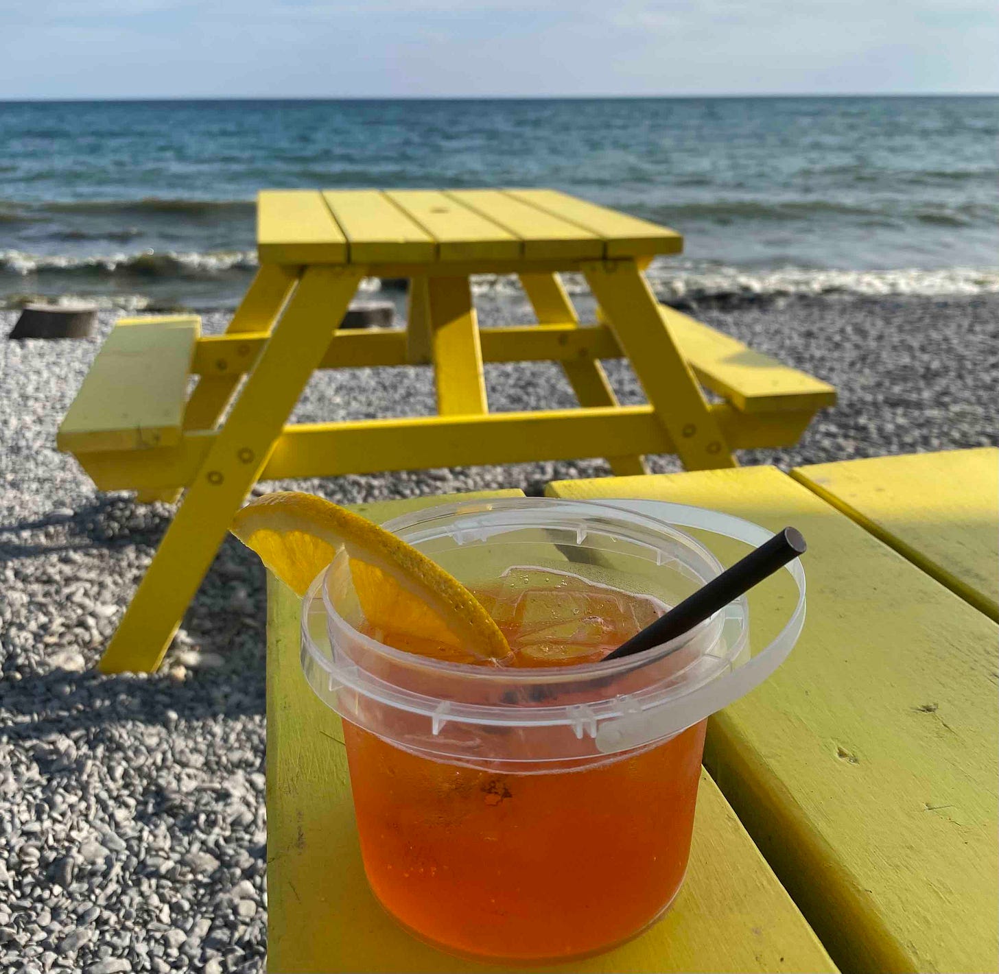 Photo of an aperol spritz in a plastic bucket on a yellow picnic table in front of a lake