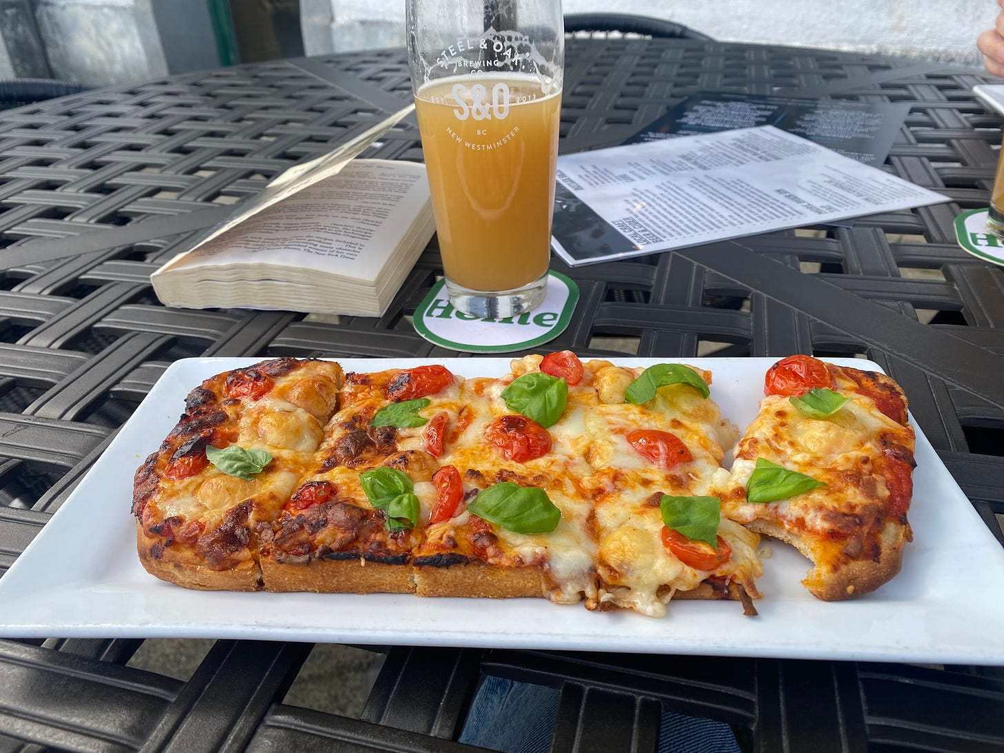 A cross-hatch patio table with a rectangular pan-style pizza on it: cheese, grape tomatoes, and basil. Behind it are a paperback book, and a glass of IPA with the Steel & Oak emblem on it.