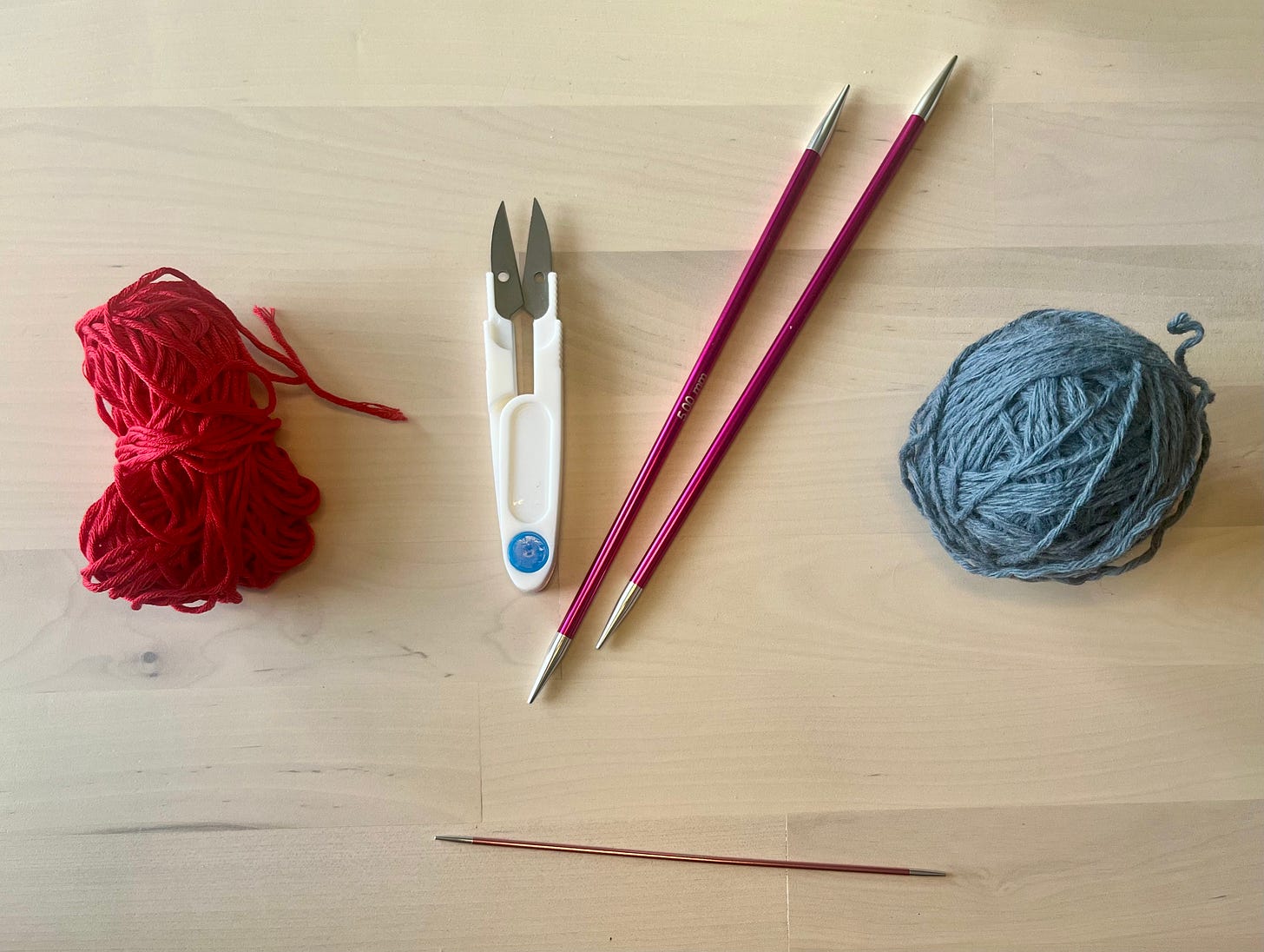 Flatlay on a wooden surface: a bundle of red waste yarn, a pair of thread snips, two magenta interchangeable needle tips, a thin double-pointed needle, and a ball of blue-grey yarn.