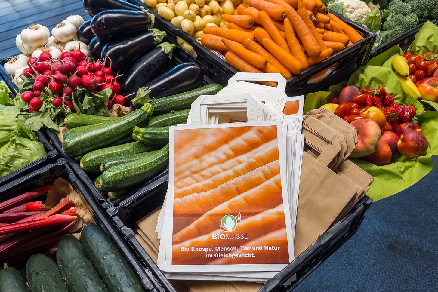 Marktstand mit einer bunten Auswahl an frischem Bio-Gemüse und Obst, darunter Zucchini, Rüebli, Radieschen, Auberginen, Broccoli, Erdbeeren und Pfirsiche. In der Mitte liegen weisse Tragetaschen mit dem orangefarbenen Bio-Suisse-Logo und dem Slogan «Bio Knospe. Mensch, Tier und Natur im Gleichgewicht.» Marktstand mit einer bunten Auswahl an frischem Bio-Gemüse und Obst, darunter Zucchini, Rüebli, Radieschen, Auberginen, Broccoli, Erdbeeren und Pfirsiche. In der Mitte liegen weisse Tragetaschen mit dem orangefarbenen Bio-Suisse-Logo und dem Slogan «Bio Knospe. Mensch, Tier und Natur im Gleichgewicht.»