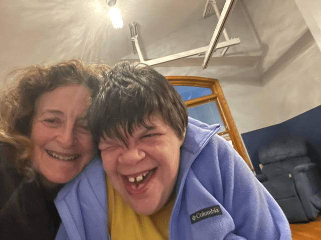 A woman and a young adult with a joyful expression, both smiling widely for a selfie, with a cozy indoor setting in the background.