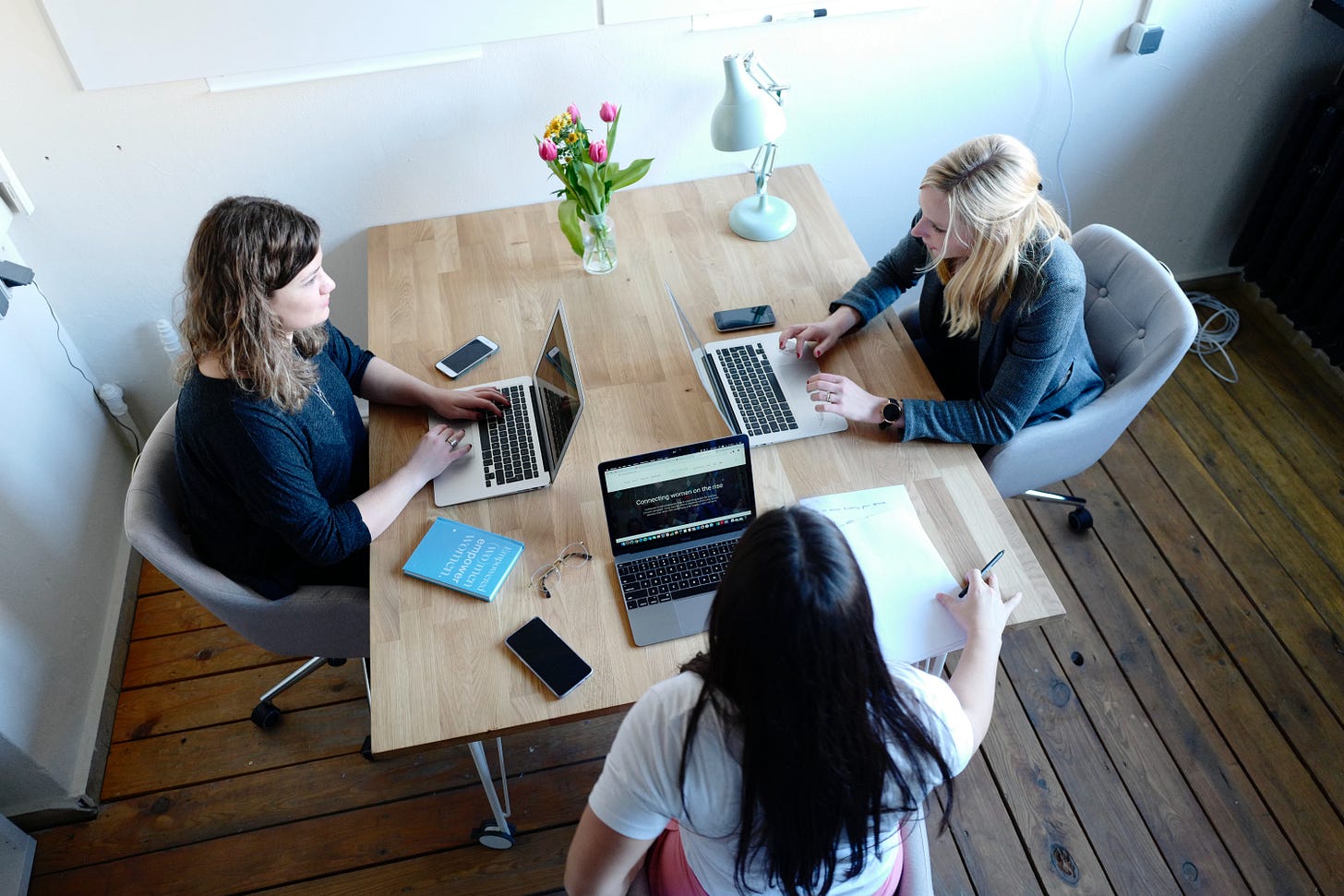 Three women work on laptops at a table with pretty flowers. Three women work on laptops at a table with pretty flowers.