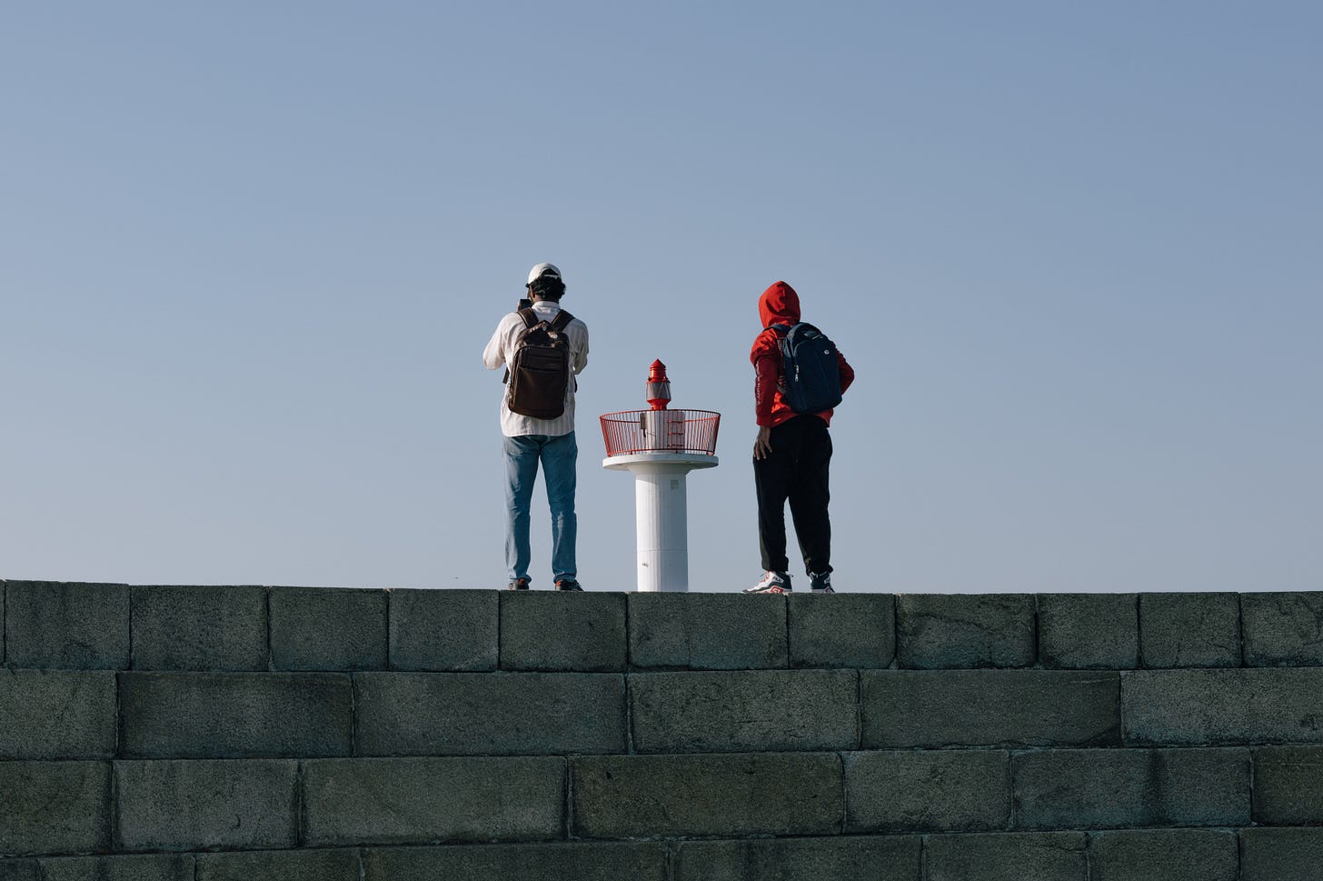 Two people with backpacks stand side by side on a pier, facing a small red navigation light against a clear blue sky. Two people with backpacks stand side by side on a pier, facing a small red navigation light against a clear blue sky.