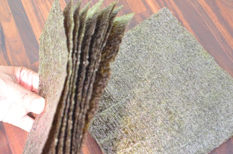 Three types of seaweed displayed side by side: a small bowl of dried mixed seaweed with thin green and dark strands, a thick glossy sheet of kombu, and a stack of crisp square sheets of nori.