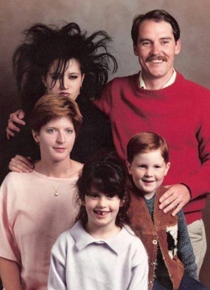 1980s era family portrait showing a Gothic teenager with dramatic teased black hair and dark makeup standing behind parents and two younger siblings, all dressed conventionally.