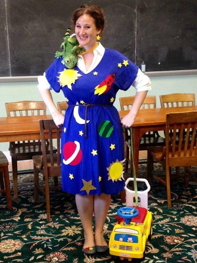 Photo of a smiling woman standing in front of a blackboard and table dressed in a Miss Frizzle outfit, with a stuffed animal lizard on her shoulder and a toddlers ride-on schoolbus by her feet