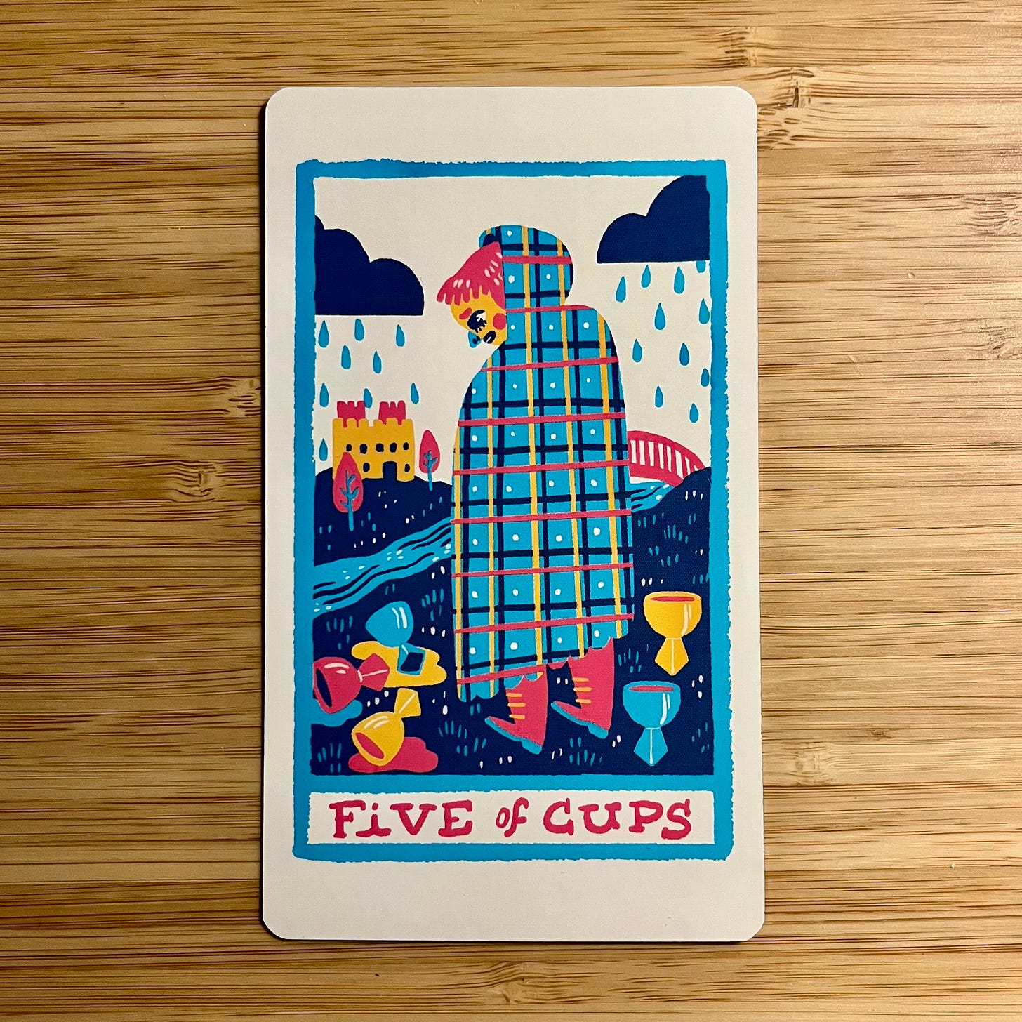 An image of a tarot card against a wood grain background. The background of the tarot card is white. The image sits within a blue outline of a vertical rectangle that has the texture of having been drawn with a crayon or oil pastel. Across the bottom of the rectangle are the words "Five of Cups" written in red capital letters. In the image stands a person with red hair, red boots, and a blue/red/yellow/black plaid hooded cloak with white polka dots. They are looking down at three spilled cups — one blue, one red, one gold — scattered across a black, grassy ground. Behind them, two cups — one blue, one gold — are still standing, liquid intact. In the mid-ground a river crosses the image. On the far right of the image there is a red bridge that leads over the river and toward a gold and red castle that sites in the far background flanked by two trees with blue trunks and red foliage. In the sky two black clouds release twenty rain drops in the shape of tears. An image of a tarot card against a wood grain background. The background of the tarot card is white. The image sits within a blue outline of a vertical rectangle that has the texture of having been drawn with a crayon or oil pastel. Across the bottom of the rectangle are the words "Five of Cups" written in red capital letters. In the image stands a person with red hair, red boots, and a blue/red/yellow/black plaid hooded cloak with white polka dots. They are looking down at three spilled cups — one blue, one red, one gold — scattered across a black, grassy ground. Behind them, two cups — one blue, one gold — are still standing, liquid intact. In the mid-ground a river crosses the image. On the far right of the image there is a red bridge that leads over the river and toward a gold and red castle that sites in the far background flanked by two trees with blue trunks and red foliage. In the sky two black clouds release twenty rain drops in the shape of tears.