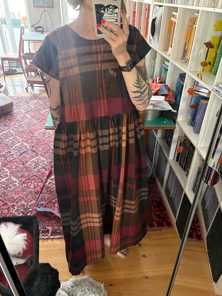 White woman with fern tattoos on her arms wears a plaid print dress with gathered waist and short sleeves in brown, cream, raspberry and pale orange. Picture on left has a loose waist with gathering free, on the right the waist is cinched in with a fabric tie belt.