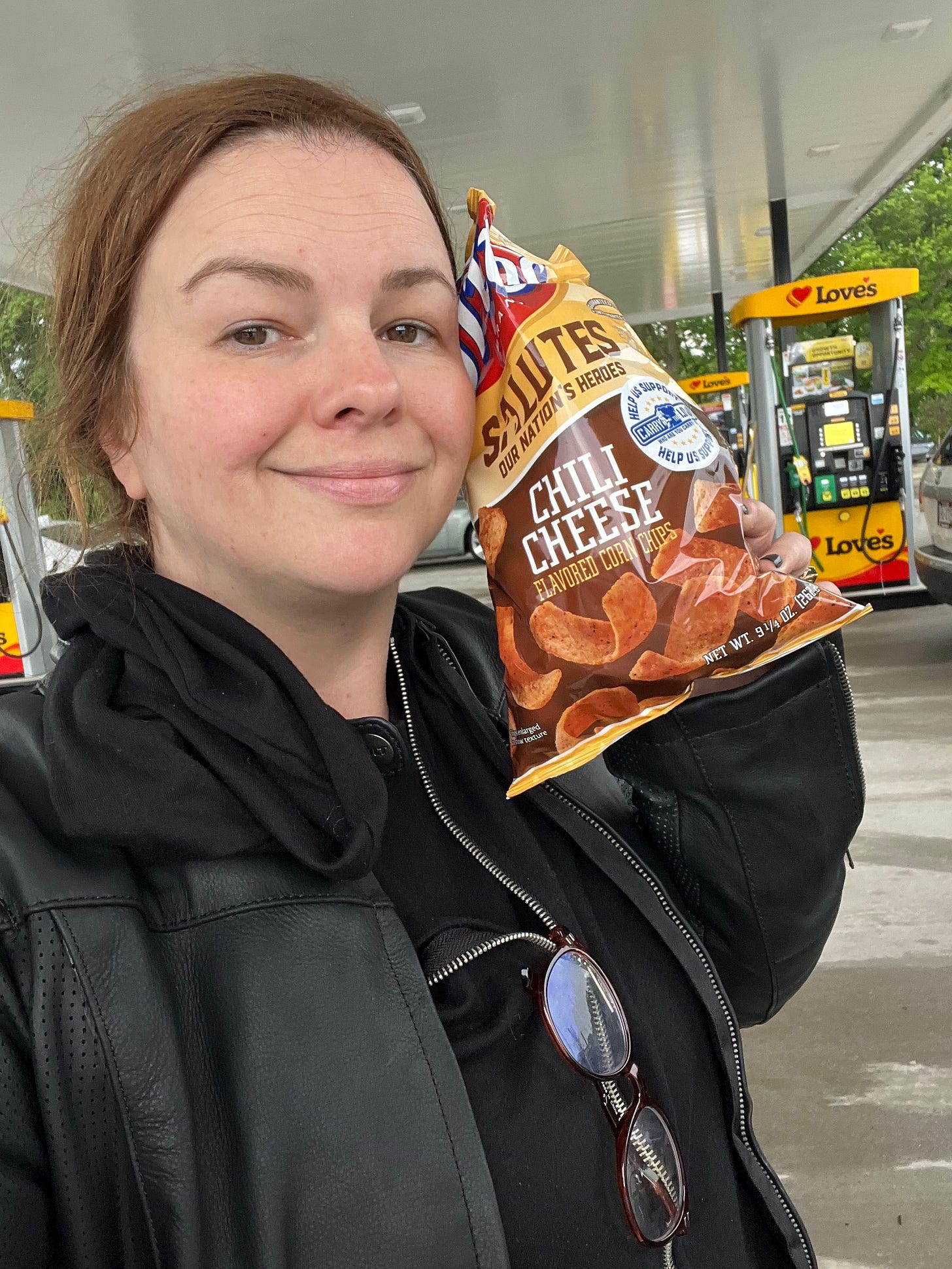 Amber takes a selfie outside of a gas station. She holds a bag of Chili Cheese Fritos next to her head and smiles. 
