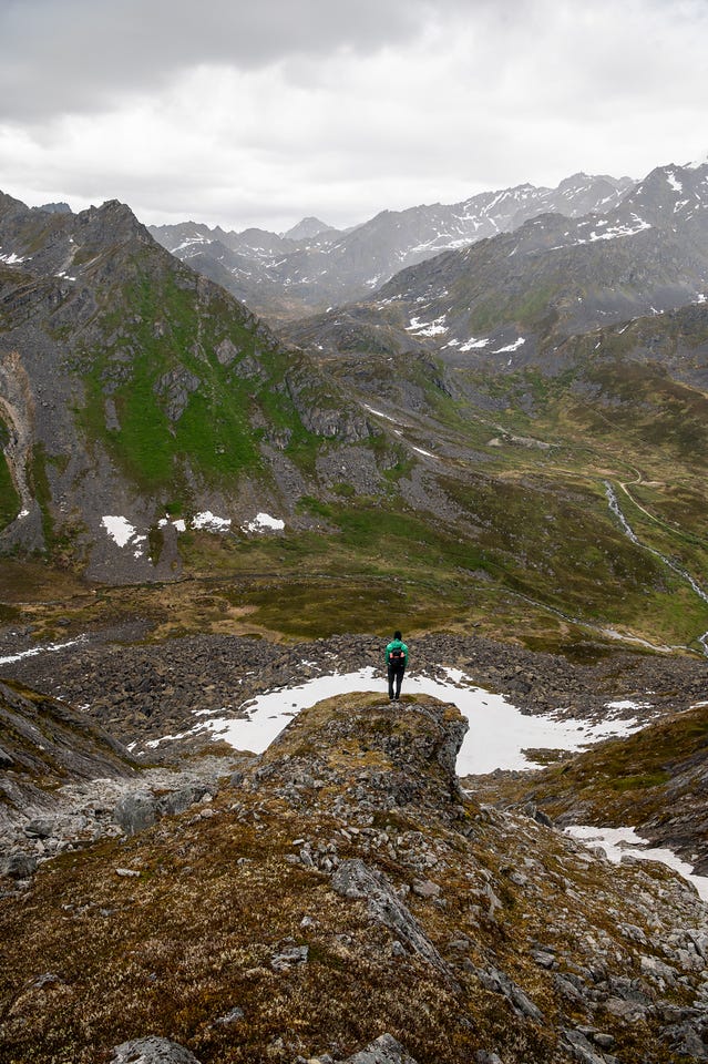 A lone hiker in a green jacket stands on a rock outcrop between two patches of old snow, back to the camera, looking down the length of a mountain valley. Green and grey slopes fall away toward a braided creek and a distant road, and ridgeline after ridgeline of dark, snow-streaked peaks recede under low grey cloud, rain hazing the farthest ranges. A lone hiker in a green jacket stands on a rock outcrop between two patches of old snow, back to the camera, looking down the length of a mountain valley. Green and grey slopes fall away toward a braided creek and a distant road, and ridgeline after ridgeline of dark, snow-streaked peaks recede under low grey cloud, rain hazing the farthest ranges.