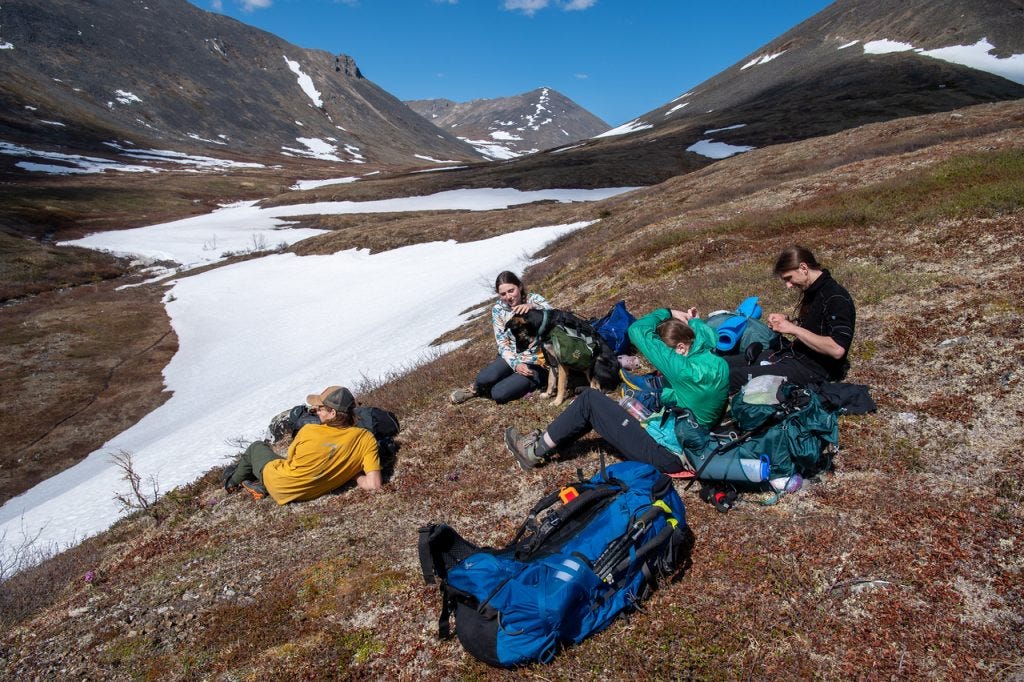 Four hikers and a dog rest on a tundra slope among scattered packs, a long ribbon of snow filling the valley below and dark peaks rising across it.