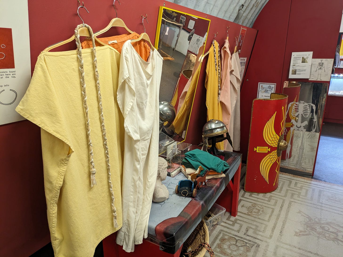 Photo of a selection of replica Roman clothes that are part of a dressing up area, including a soldier's helmet, a shield, and some simple tunics