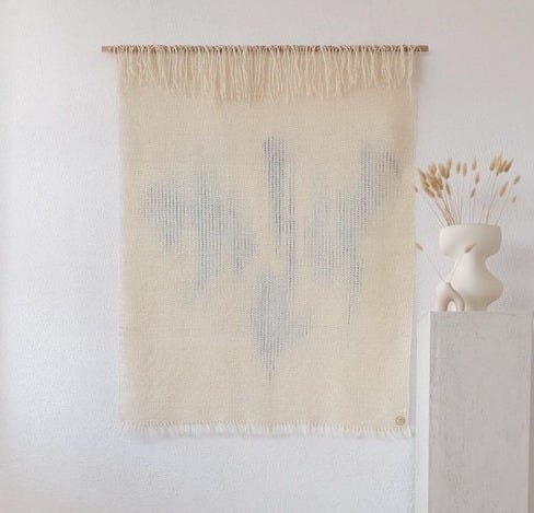May include: A white woven wall hanging with a blue abstract design hangs on a white wall. A white pedestal with a white ceramic vase with dried beige grasses sits in front of the wall hanging.