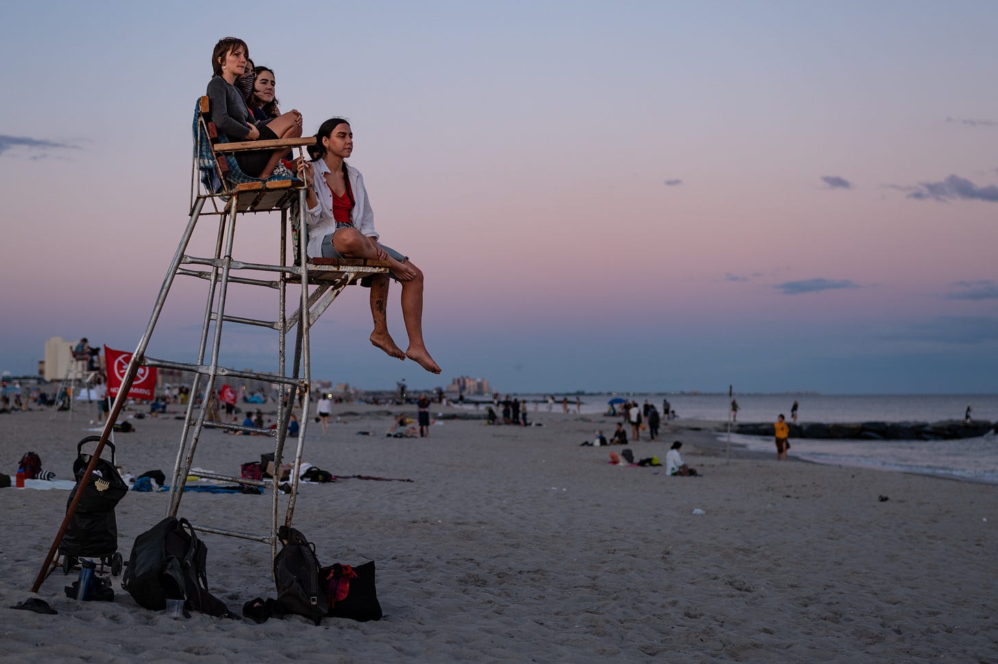 Three young women sitting on a wooden lifeguard chair on the sand at dusk at Rockaway Beach, Queens, watching a public dance, with a pink and blue sky over the ocean.