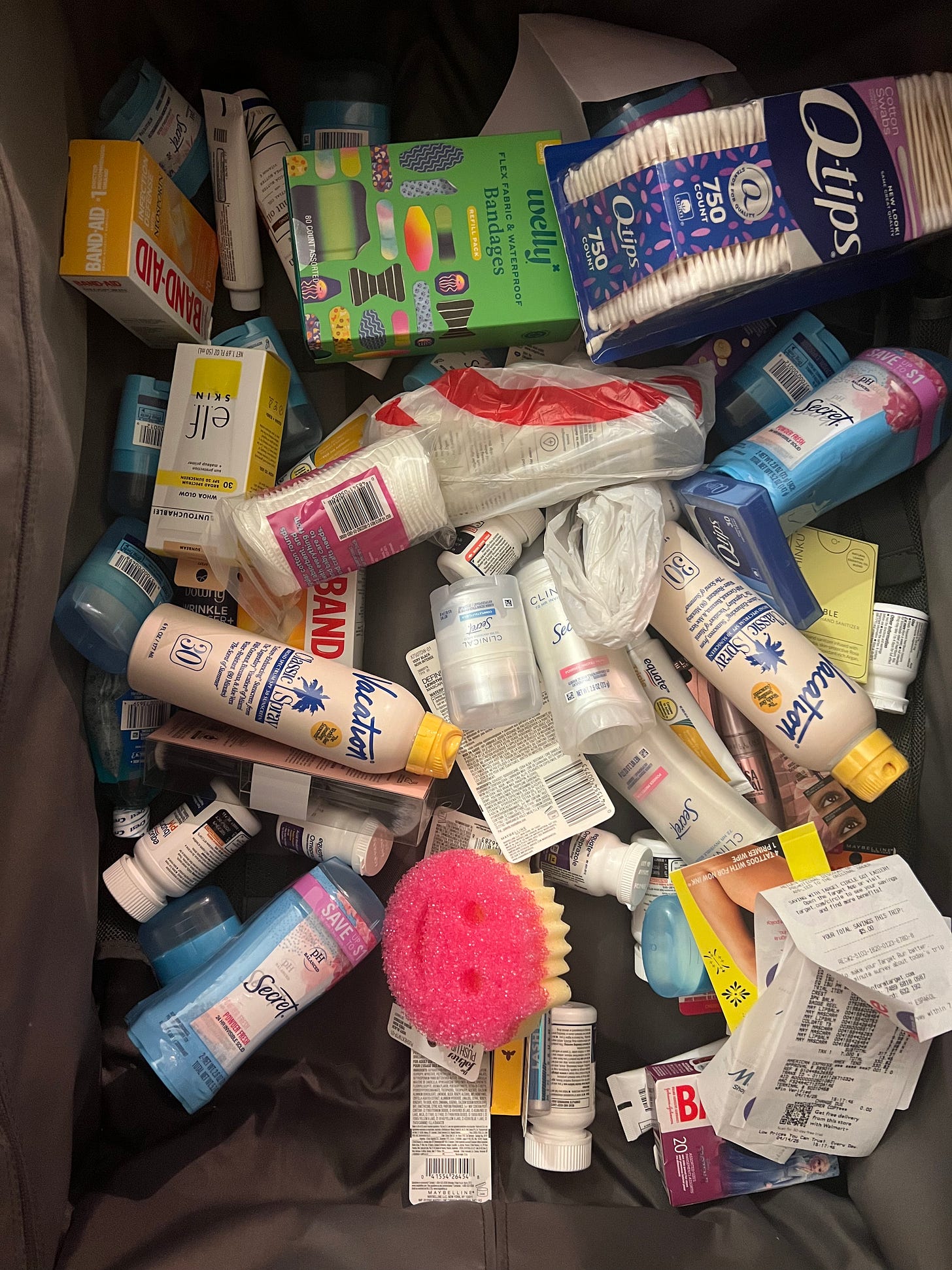 A flat-lay view inside a gray suitcase overflowing with a US drugstore beauty haul. Visible products include Vacation Classic Spray sunscreen, several sticks of Secret deodorant, Welly bandages, Band-Aid boxes, e.l.f. Skin products, and cotton swabs, all scattered alongside a store receipt. A flat-lay view inside a gray suitcase overflowing with a US drugstore beauty haul. Visible products include Vacation Classic Spray sunscreen, several sticks of Secret deodorant, Welly bandages, Band-Aid boxes, e.l.f. Skin products, and cotton swabs, all scattered alongside a store receipt.