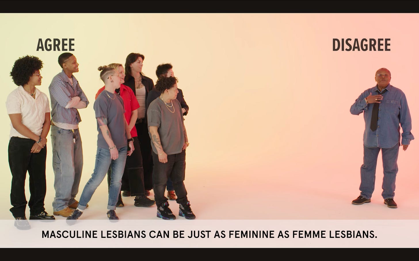 Screenshot of the Youtube video, “Will 8 Masc Lesbians Disagree With Each Other?” A prompt at the bottom of the screen says, “Masculine lesbians can be just as feminine as feminine lesbians”. A group of lesbians stand on one side of the room agreeing with the prompt, while one elderly black butch lesbian stands on the other side, disagreeing. 
