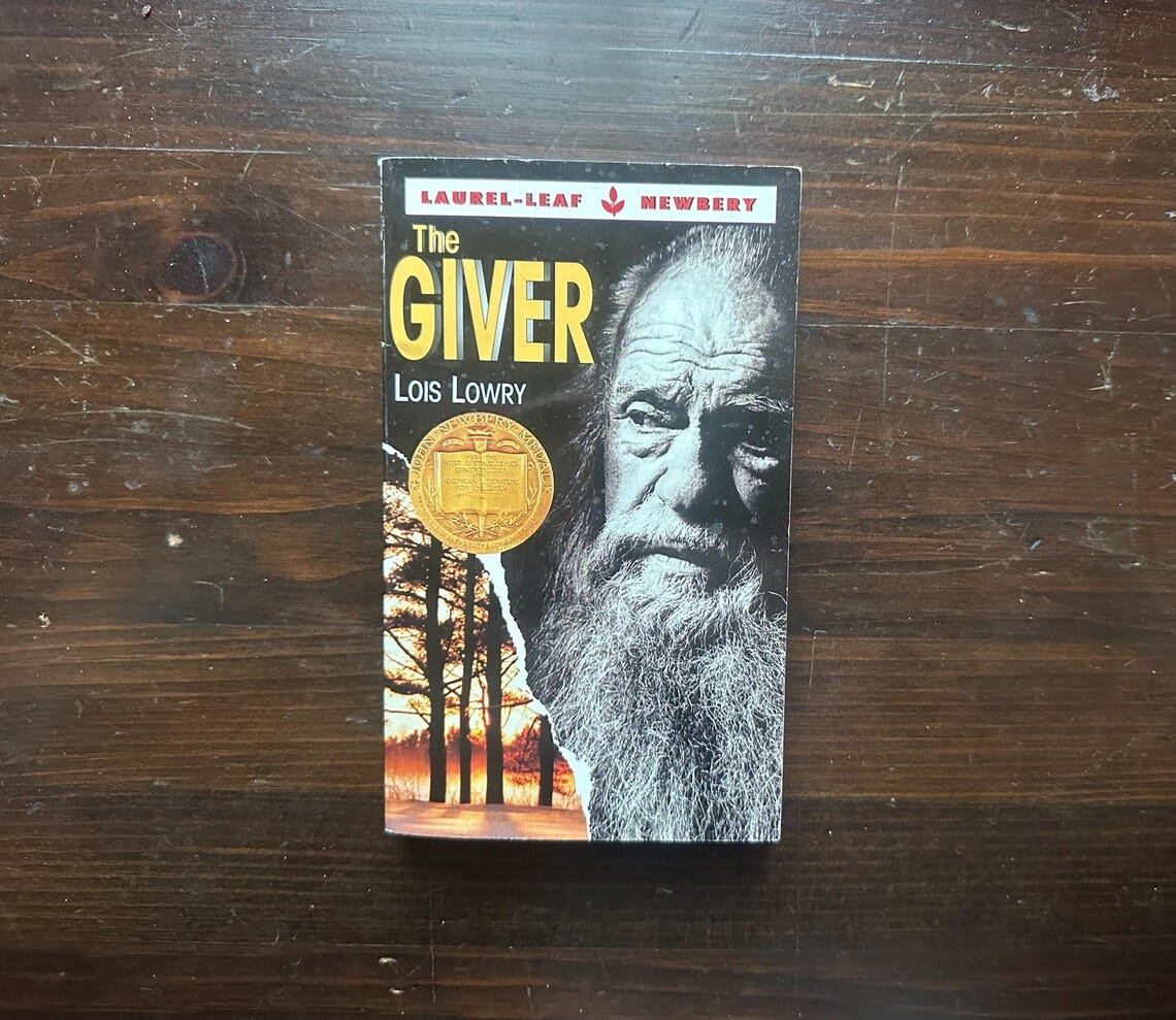 May include: The Giver by Lois Lowry, a young adult novel with a black and white cover featuring a man with a long white beard and a gold medal. May include: The Giver by Lois Lowry, a young adult novel with a black and white cover featuring a man with a long white beard and a gold medal.