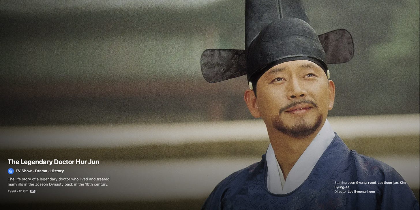 A screenshot of the Apple TV listing for The Legendary Doctor Hur Jun. A man in Joseon-era clothing and a traditional black hat looks upward slightly. On-screen text shows the title, genre (“TV Show · Drama · History”), and a brief description of the series. A screenshot of the Apple TV listing for The Legendary Doctor Hur Jun. A man in Joseon-era clothing and a traditional black hat looks upward slightly. On-screen text shows the title, genre (“TV Show · Drama · History”), and a brief description of the series.