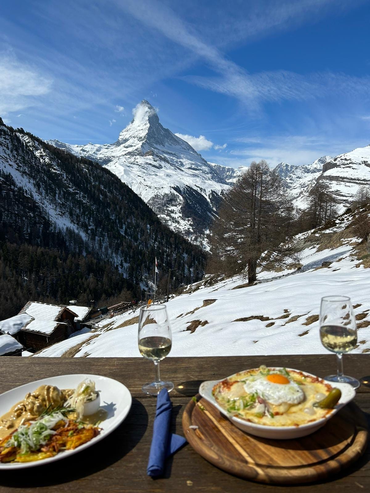 A winter photo of the matterhorn with two plates of traditional swiss food and two glasses of white wine.