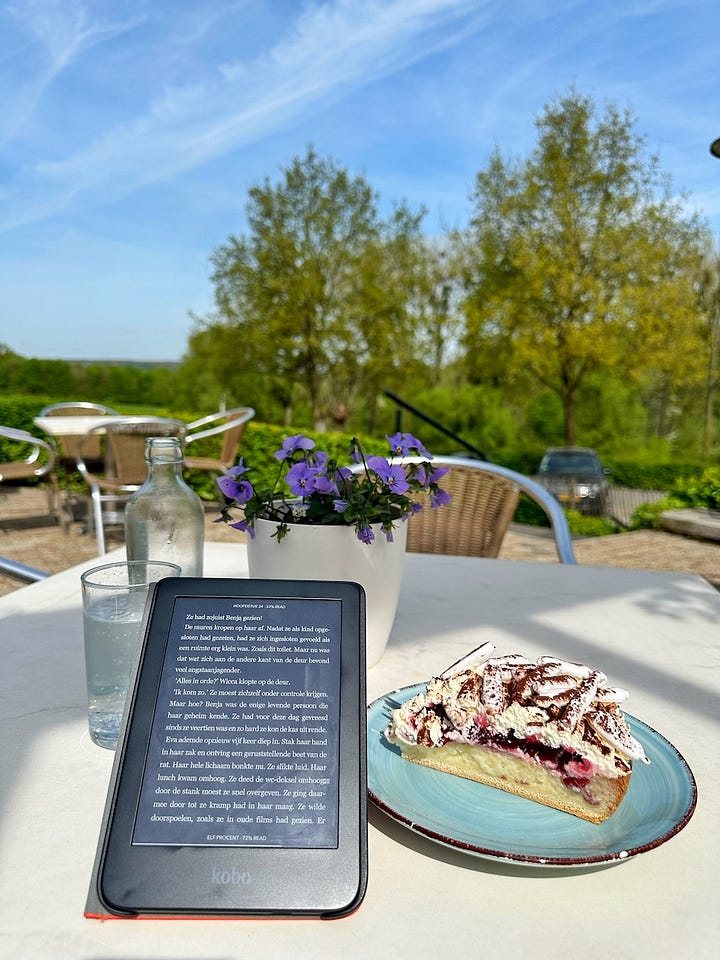 Een e-reader en een stuk vlaai op een terrastafel en een oranje e-readerhoes op een wit dekbed in een halfdonkere kamer.