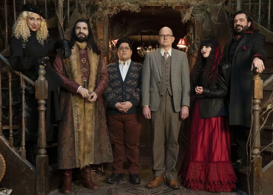In the entryway of a very old and derelict house, at the base of a grand dual staircase, stands a line of six eccentric individuals facing forward. The two in the middle are serious-looking men, one pale and bald in a suit, the other a dark-haired shorter man in a cardigan sweater and brown pants. The other four individuals, a man and woman on each side, are all vampires smiling in eclectic outfits from various eras and regions of the world. 