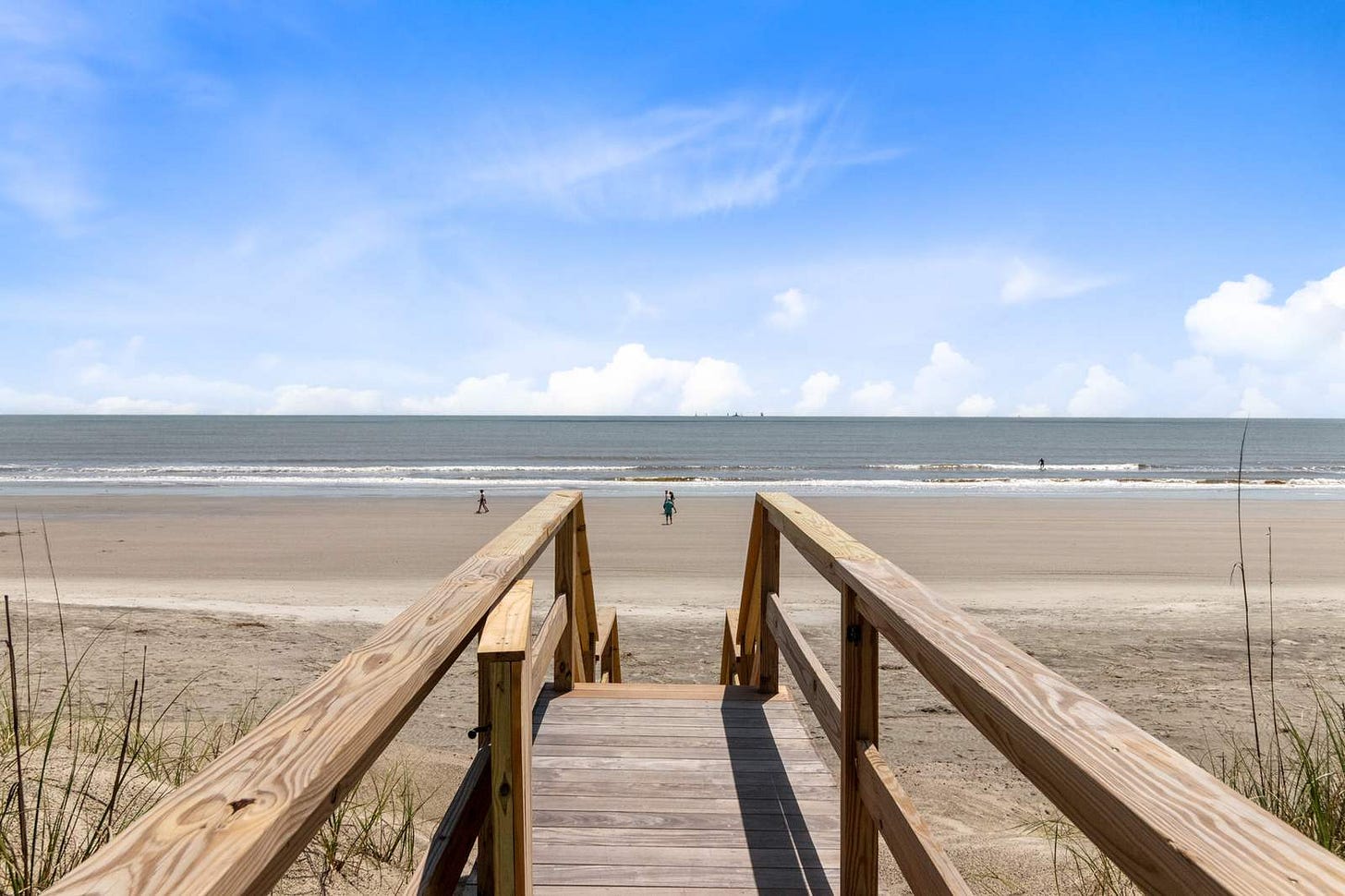 A wooden beach boardwalk leading through dune grass down to wide flat sand, with the ocean and a bright blue sky beyond and two people walking in the distance. A wooden beach boardwalk leading through dune grass down to wide flat sand, with the ocean and a bright blue sky beyond and two people walking in the distance.