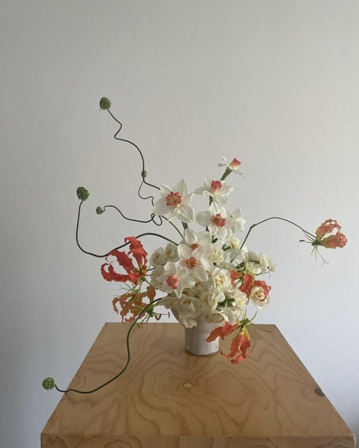 This may contain: an arrangement of flowers in a vase on a wooden table with white walls behind it