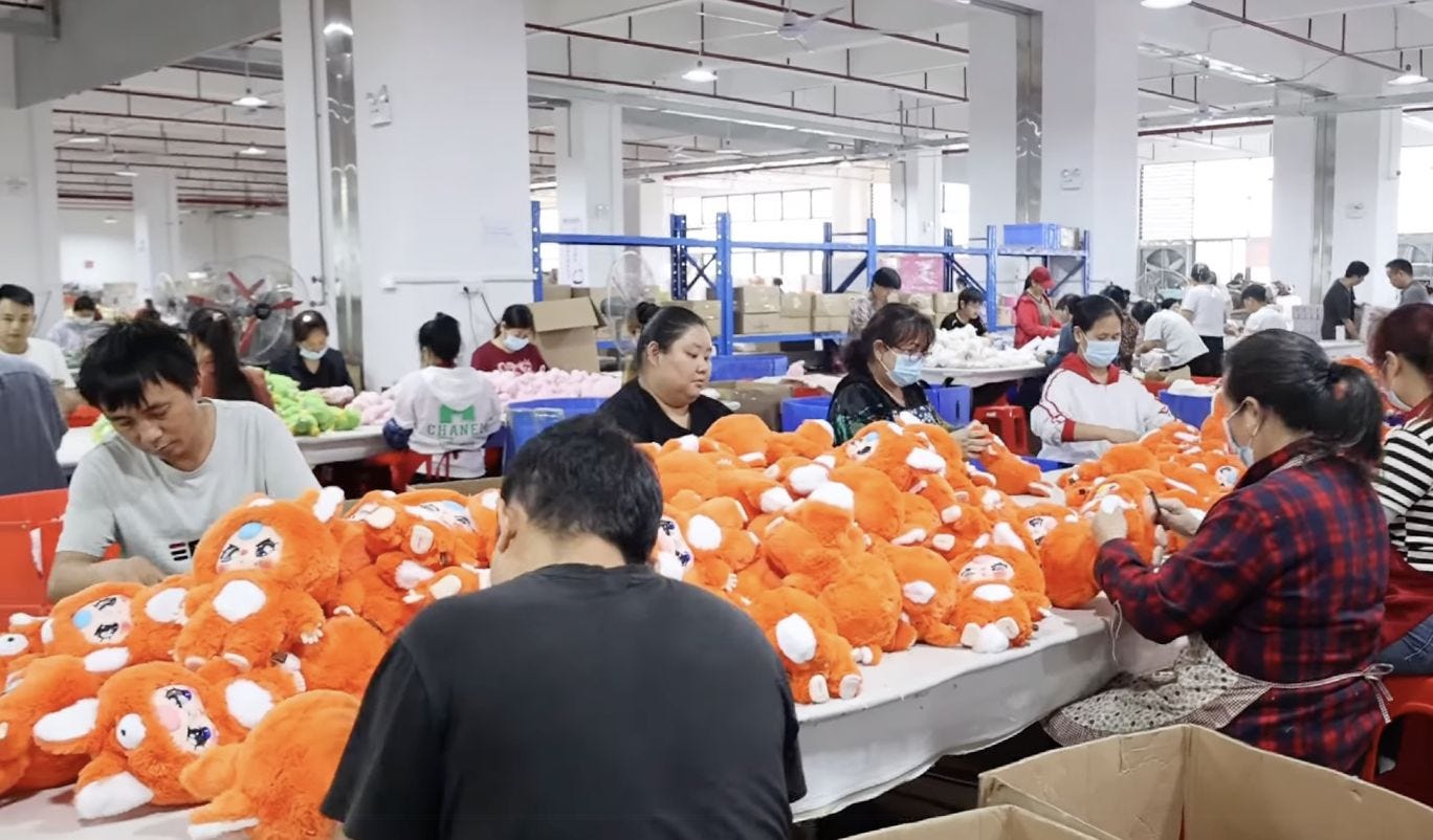 Wide shot of a plush-toy factory: rows of workers—some wearing masks—sit around tables piled high with vivid orange Baby Three dolls, stitching and assembling them amid white pillars and industrial shelves. Wide shot of a plush-toy factory: rows of workers—some wearing masks—sit around tables piled high with vivid orange Baby Three dolls, stitching and assembling them amid white pillars and industrial shelves.