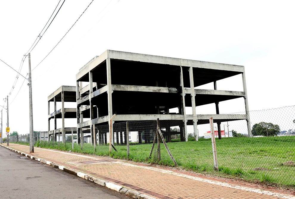 Estrutura de concreto inacabada com quatro níveis, cercada por cerca de metal galvanizado, situada em terreno com grama, ladeada por via pavimentada com tijolos, poste de energia com fios elétricos, áreas verdes ao fundo e céu nublado. Estrutura de concreto inacabada com quatro níveis, cercada por cerca de metal galvanizado, situada em terreno com grama, ladeada por via pavimentada com tijolos, poste de energia com fios elétricos, áreas verdes ao fundo e céu nublado.