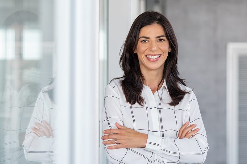 A professional woman smiling confidently while standing with her arms crossed. She has long dark hair and is wearing a white blouse with a black grid pattern. The background shows a modern office space with glass walls, providing a clean and contemporary setting. A professional woman smiling confidently while standing with her arms crossed. She has long dark hair and is wearing a white blouse with a black grid pattern. The background shows a modern office space with glass walls, providing a clean and contemporary setting.