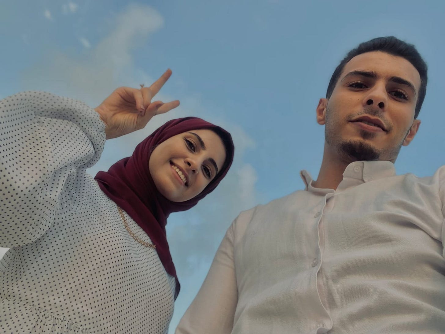 Two people are photographed from a low angle against a blue sky with light clouds. One person on the left is smiling and wearing a maroon headscarf and a light-colored, long-sleeved top with small dark polka dots, holding up a peace sign with one hand. The person on the right is wearing a light-colored button-up shirt and looking down toward the camera with a neutral expression. Two people are photographed from a low angle against a blue sky with light clouds. One person on the left is smiling and wearing a maroon headscarf and a light-colored, long-sleeved top with small dark polka dots, holding up a peace sign with one hand. The person on the right is wearing a light-colored button-up shirt and looking down toward the camera with a neutral expression.