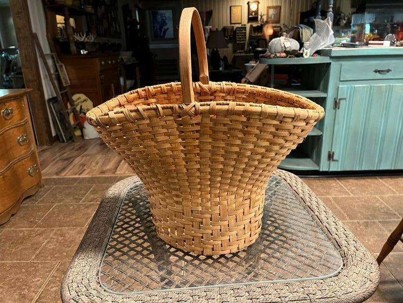 May include: A light brown, woven, handcrafted basket with a handle. The basket is wide at the top and tapers down to a smaller base. It's made of light-colored wicker or similar material, creating a rustic and vintage aesthetic. The basket's construction shows a tightly woven pattern, suggesting durability and quality craftsmanship.  The handle is seamlessly integrated into the basket's structure, adding to its overall design. This type of basket is ideal for storage, display, or as a decorative element in a home or shop setting.