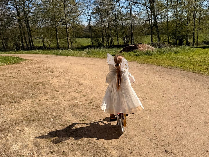 Four photos of the château grounds: a grassy track running along the edge of a sunlit wood, a girl in a pink fairy costume heading up a forest path, a girl in a white fairy dress riding a balance bike down a gravel drive, and a wicker bicycle basket holding two fresh eggs resting on leaves.